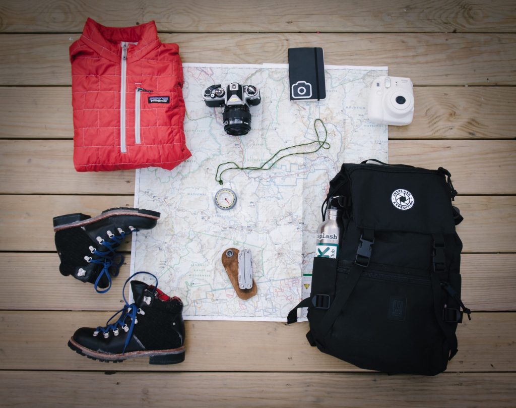 back pack, walking shows, map, camera and coat, on a wooden floor.